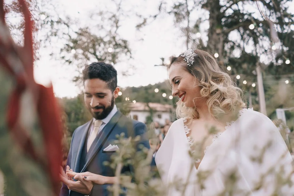 Casal de noivos sorrindo em cerimônia ao ar livre decorada com luzes no fundo.