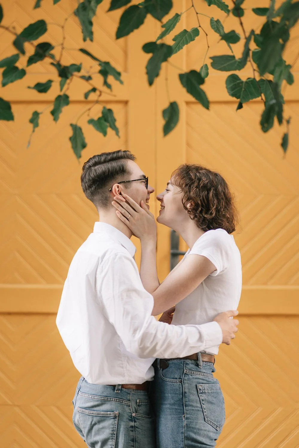 Um casal apaixonado se beijando, com a mulher segurando o rosto do homem, em um ambiente com parede de madeira e folhas verdes penduradas.