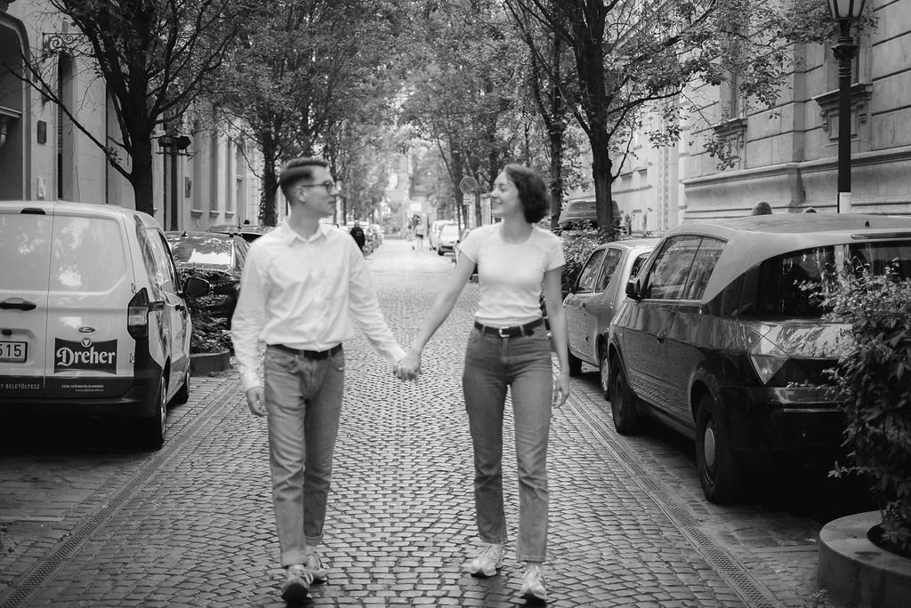 Casal andando de mãos dadas na rua de paralelepípedos, cercado por carros estacionados e árvores ao lado de edifícios.