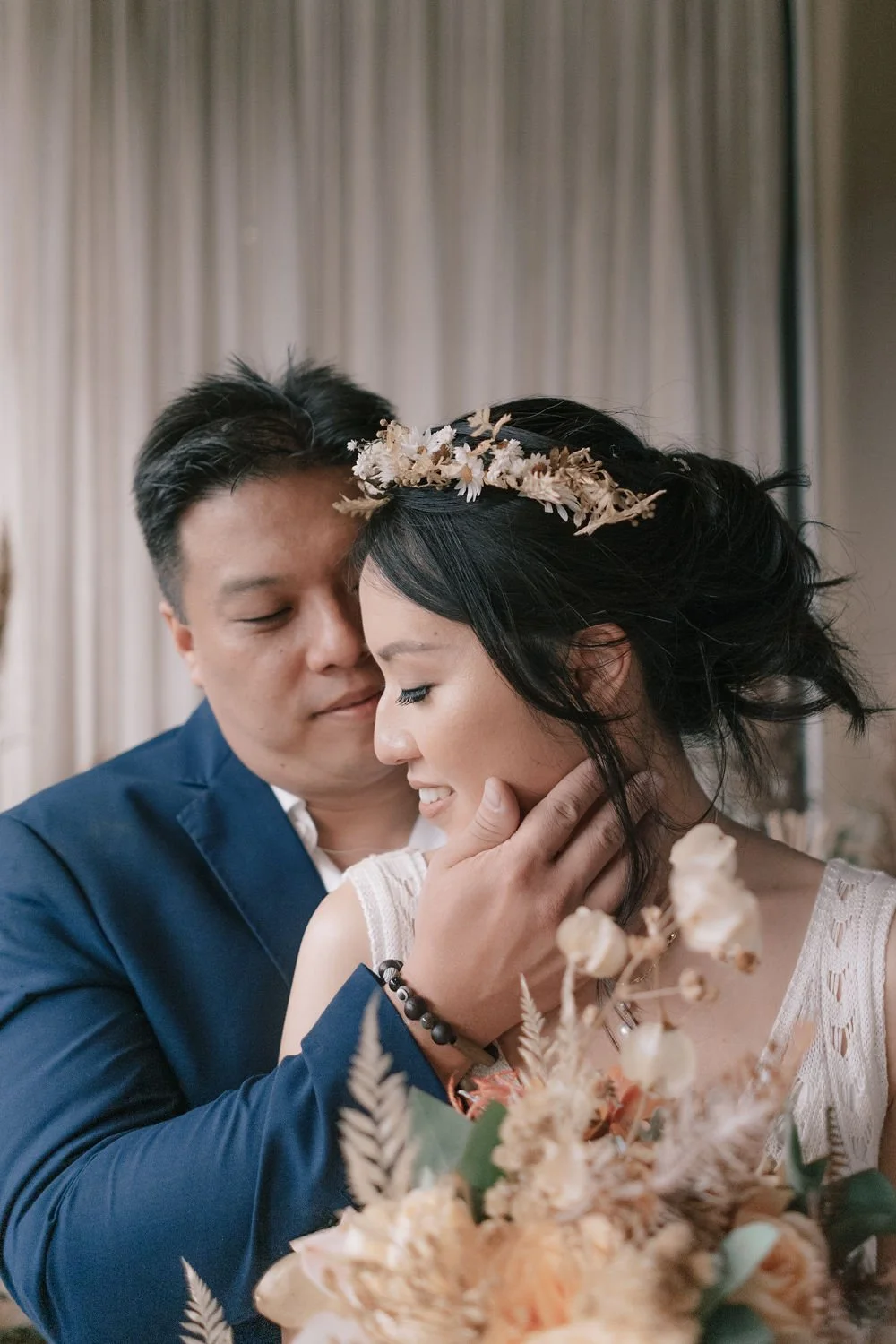 Casal apaixonado em momento romântico, com o homem segurando o rosto da mulher enquanto eles se beijam, decorado com flores e cortinas ao fundo.