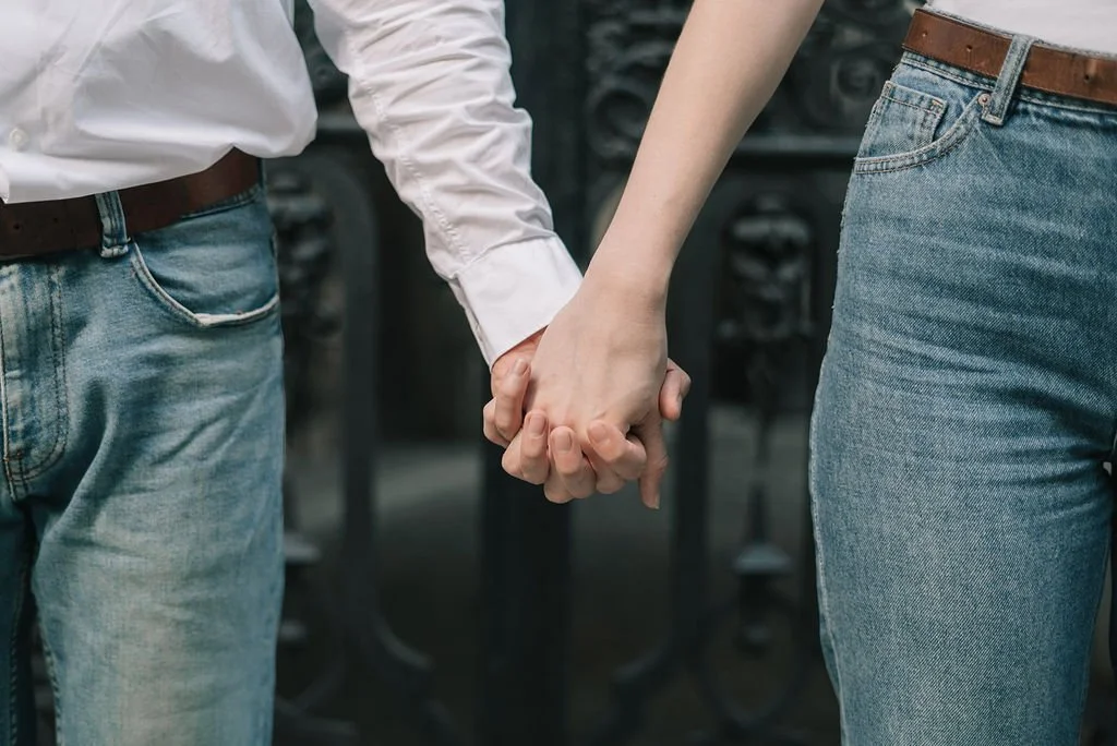 Pessoas segurando as mãos entre si, com roupas casuais, incluindo jeans e uma camisa branca, em um ambiente interno