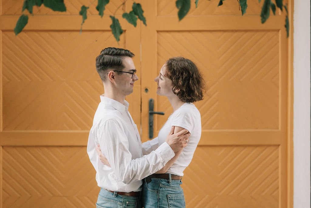 Um casal tocando as mãos um no outro frente a uma porta de madeira, sorrindo um para o outro.