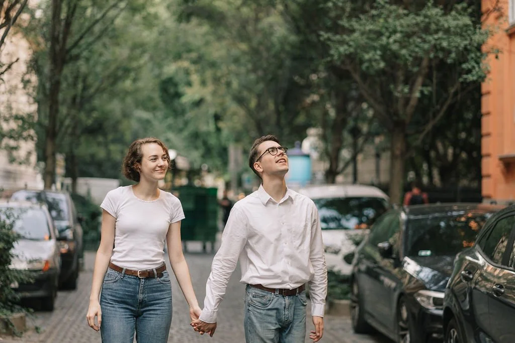 Casal caminhando de mãos dadas na rua com árvores e carros ao redor.