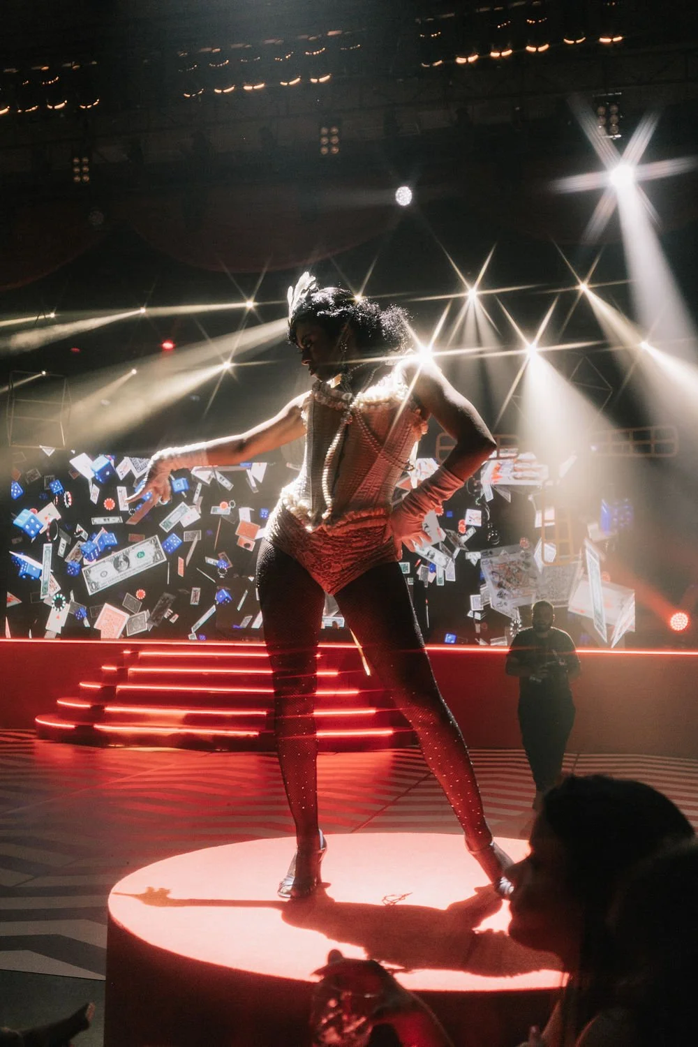 Performista em palco com iluminação intensa, roupas brilhantes, dançando e com dinheiro de mentira voando ao redor, em ambiente de show ou evento.