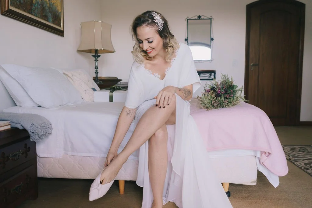 Mulher sorrindo sentada na cama, vestindo um robe branco, colocando um sapato de salto em um dos pés.