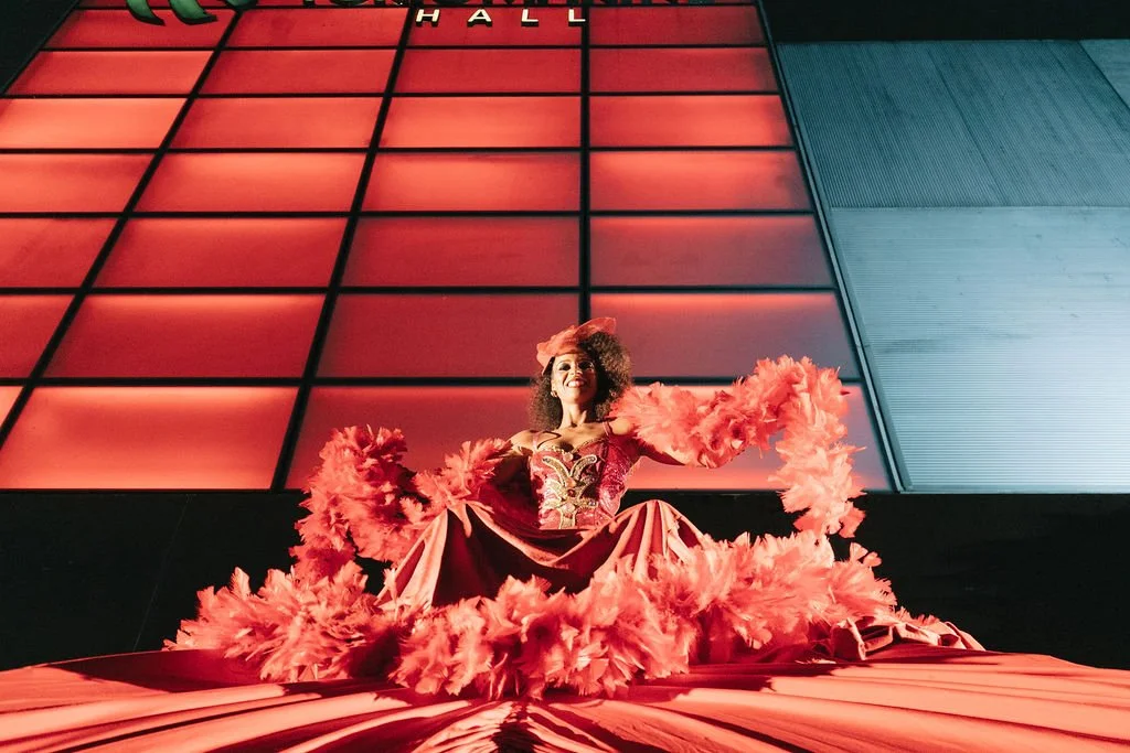 Mulher vestida com traje vermelho e frufrus, sorrindo, em palco com fundo de painel iluminado vermelho e azul.