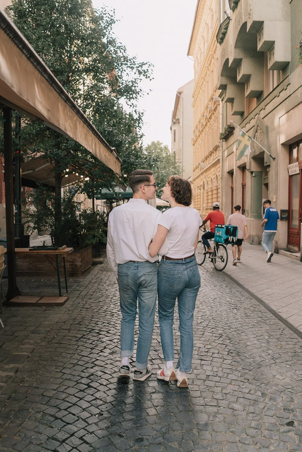 Casal caminhando de mãos dadas por rua de paralelepípedos, com prédios antigos ao fundo e pessoas ao redor, incluindo um ciclista.