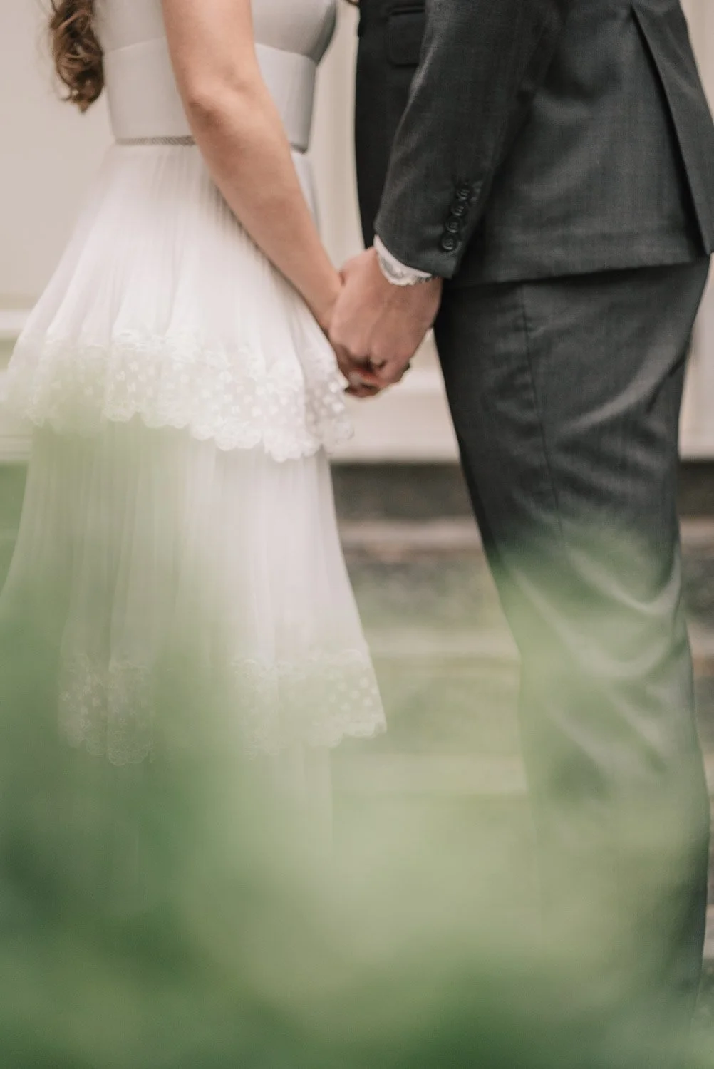 Casal de casamento segurando as mãos, focando na roupa e nas mãos deles, com o vestido de noiva branco e o terno escuro.