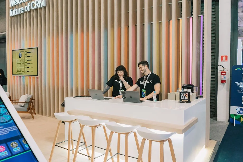 Duas pessoas trabalhando em um balcão de recepção de evento ou feira, com fundo decorativo de painéis de madeira coloridos, usando camisetas pretas com o logo 'FastBack'.
