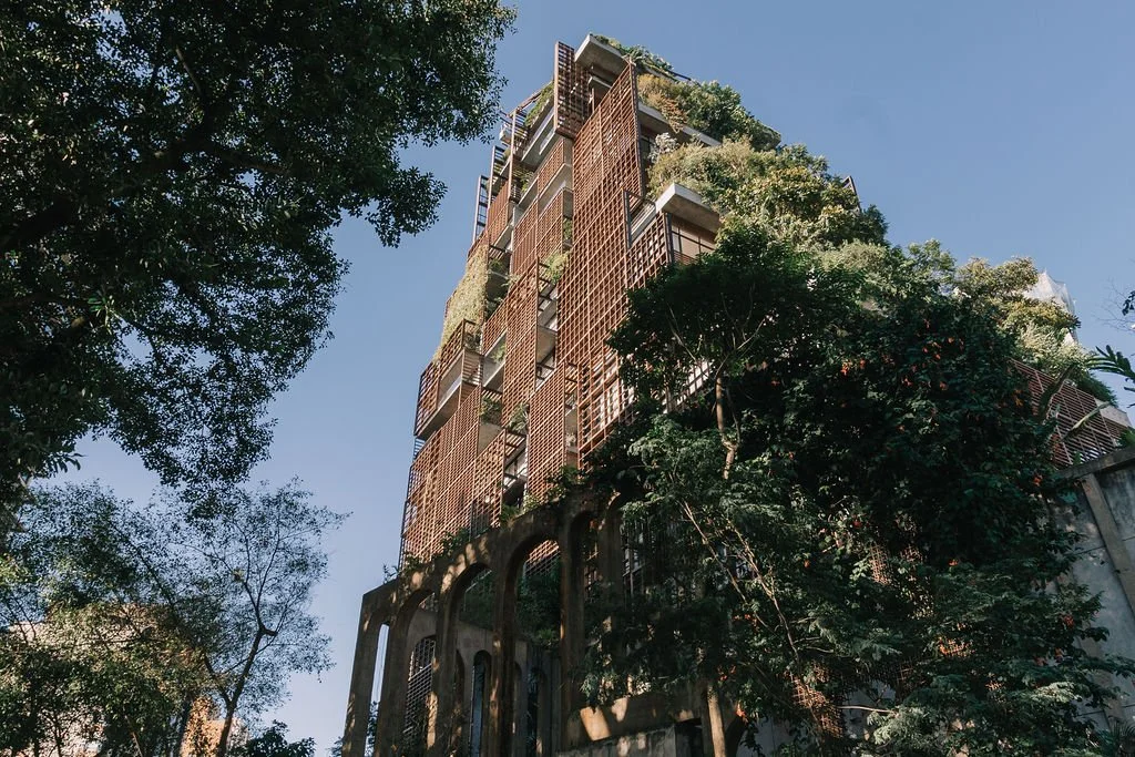 Prédio moderno com muitos jardins no exterior, cercado por árvores verdes, visto de baixo para cima contra um céu azul.