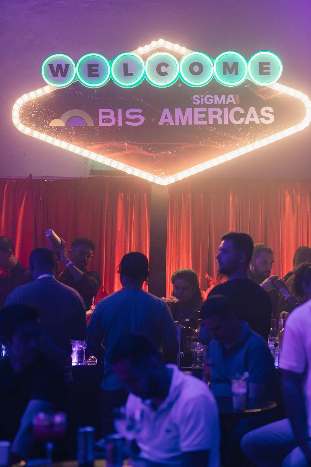 Evento com uma placa de neon dizendo 'Welcome' e 'BIS Americas', com muitas pessoas entre elas jovens, alguns segurando bebidas, em um ambiente com cortinas vermelhas.