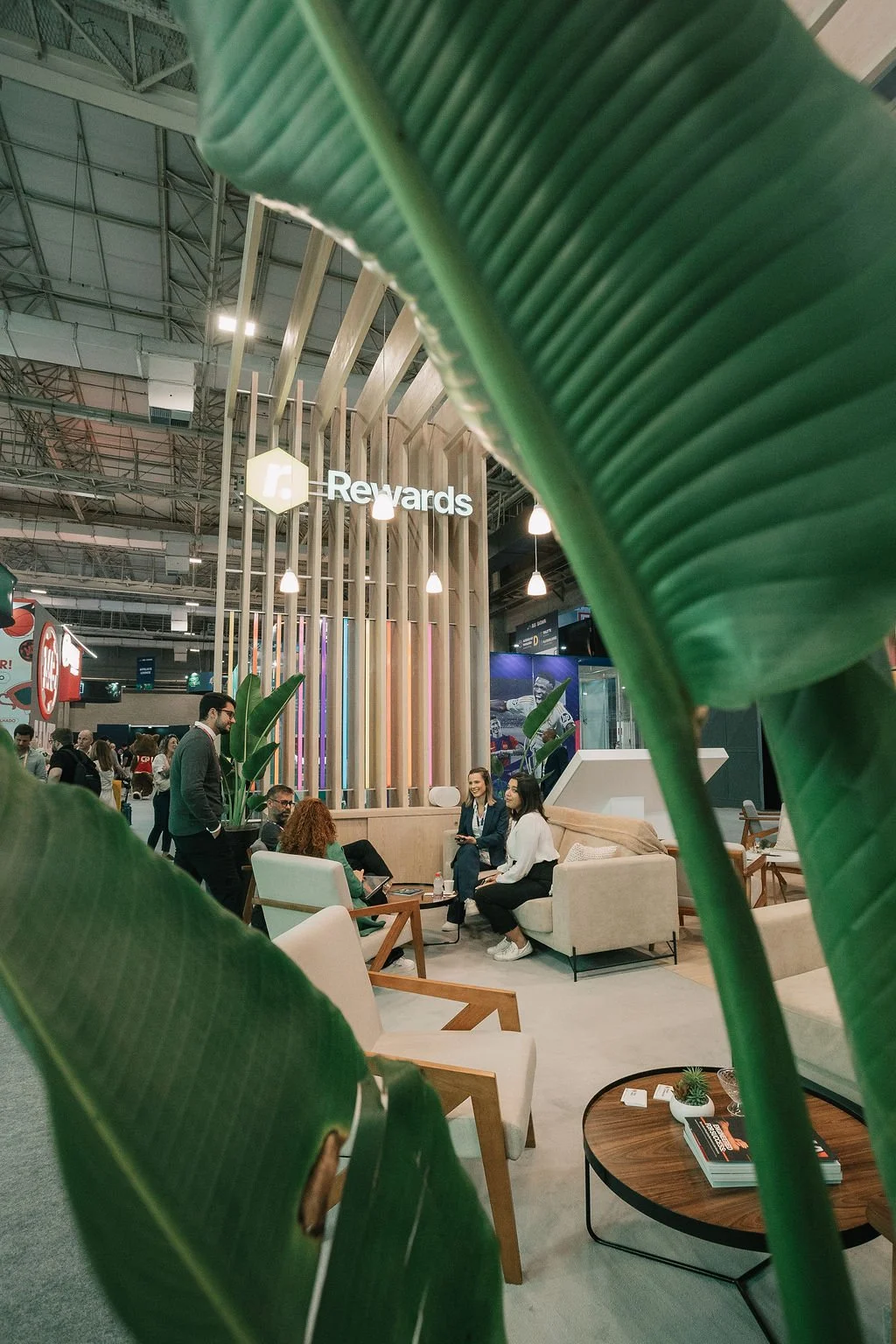 Área de lounge com pessoas conversando, móveis confortáveis e decoração moderna, vista parcialmente obstruída por folhas verdes.