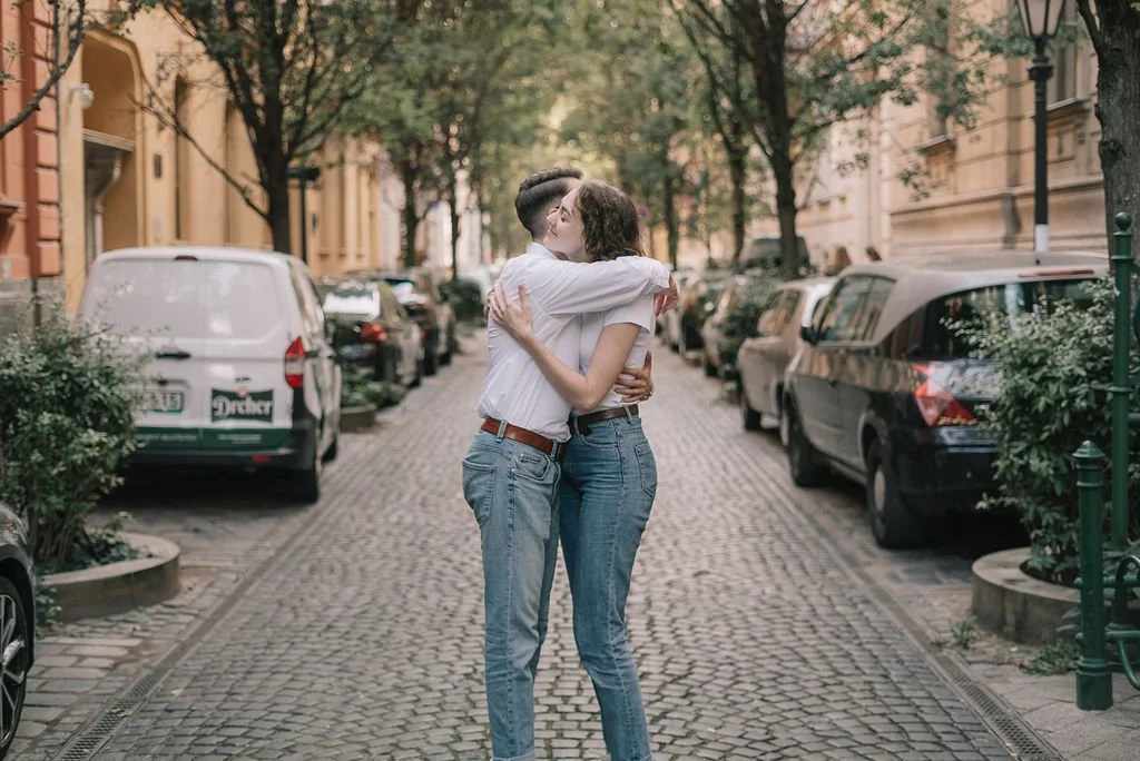 Casal abraçado no meio de uma rua de paralelepípedos com árvores e carros estacionados.