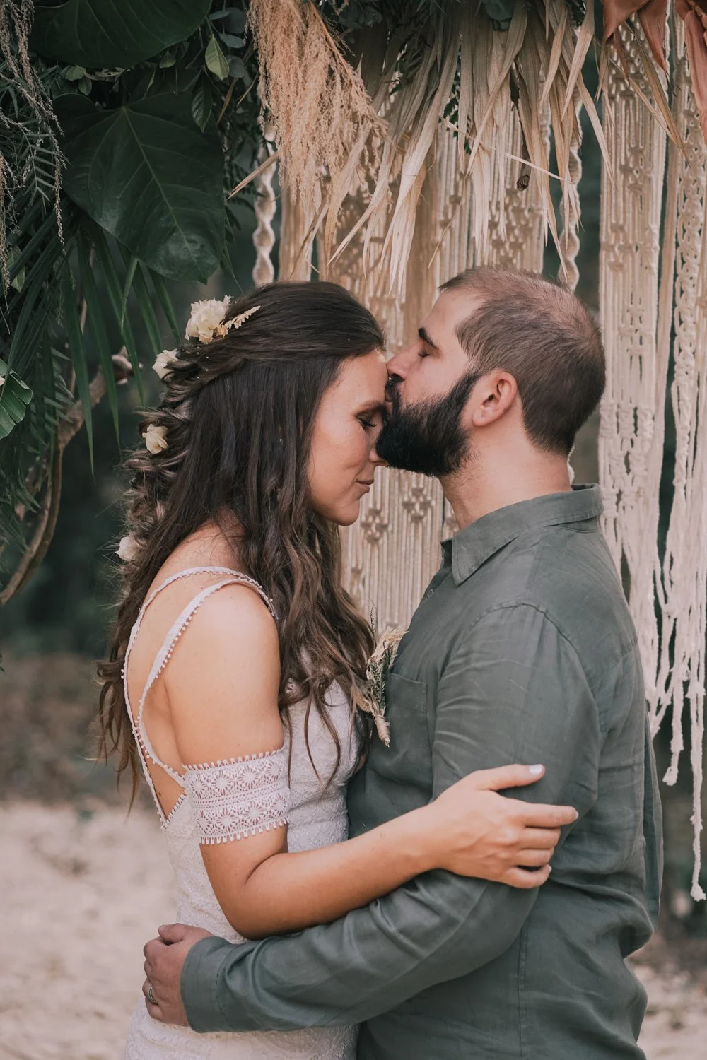 Casal se beijando com os olhos fechados, em cerimônia ao ar livre, cercados de plantas verdes e decoração de crochê.
