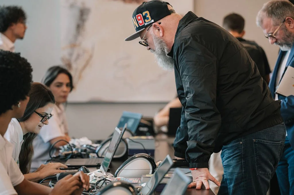 Homem de cabelo e barba grisalhos usando boné e jaqueta preta, inclinado sobre uma mesa de trabalho com computadores, cercado por várias pessoas em uma sala de reunião.