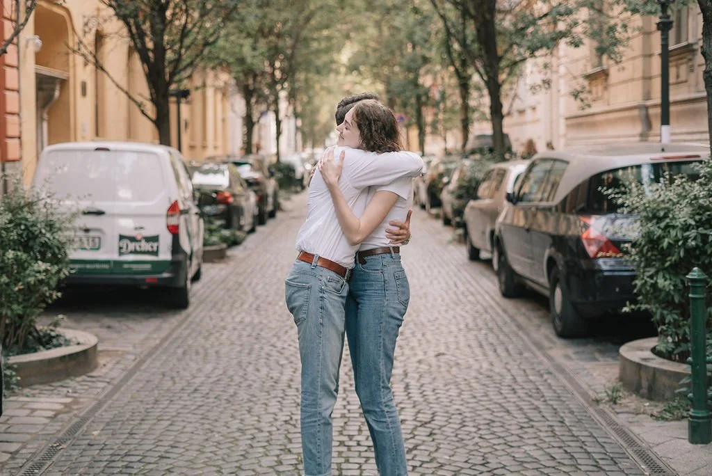 Duas pessoas se abraçando na rua de paralelepípedo, com carros estacionados ao lado e árvores ao fundo.