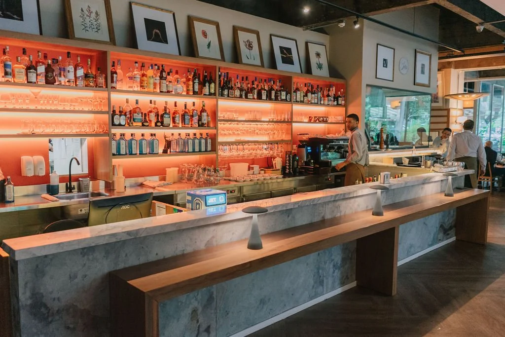 Bar com várias garrafas de bebidas alcoólicas, espelhos, quadros decorativos na parede, computadores e pessoas ao fundo, com ambiente iluminado e janelas grandes.