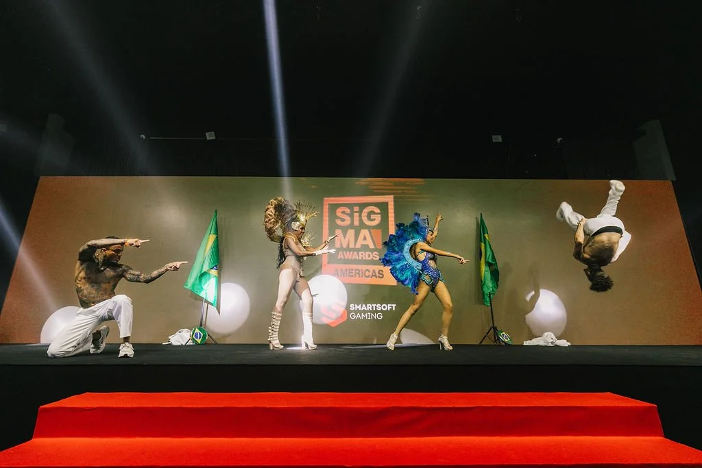 Quatro pessoas em roupas de dança apresentando na fase de um evento com fundo verde e dois um pavilhão do Brasil e a logo do evento SIGMA Awards.