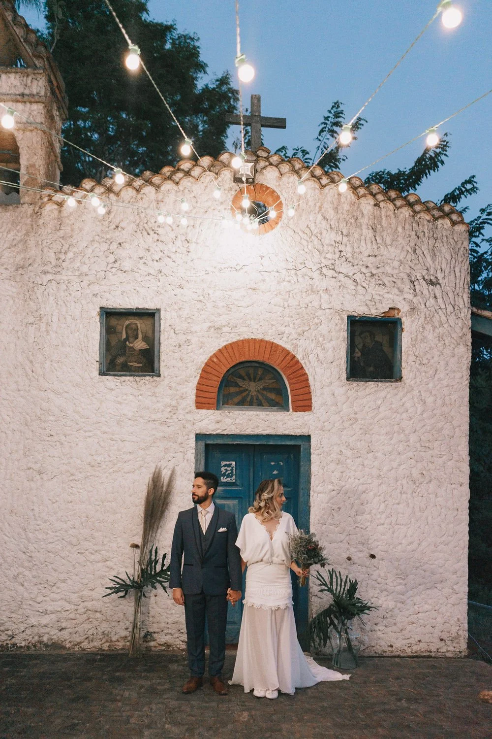 Casal de casamento segurando as mãos diante de uma igreja de paredes brancas, com decoração de plantas e luzes suspensas ao entardecer.