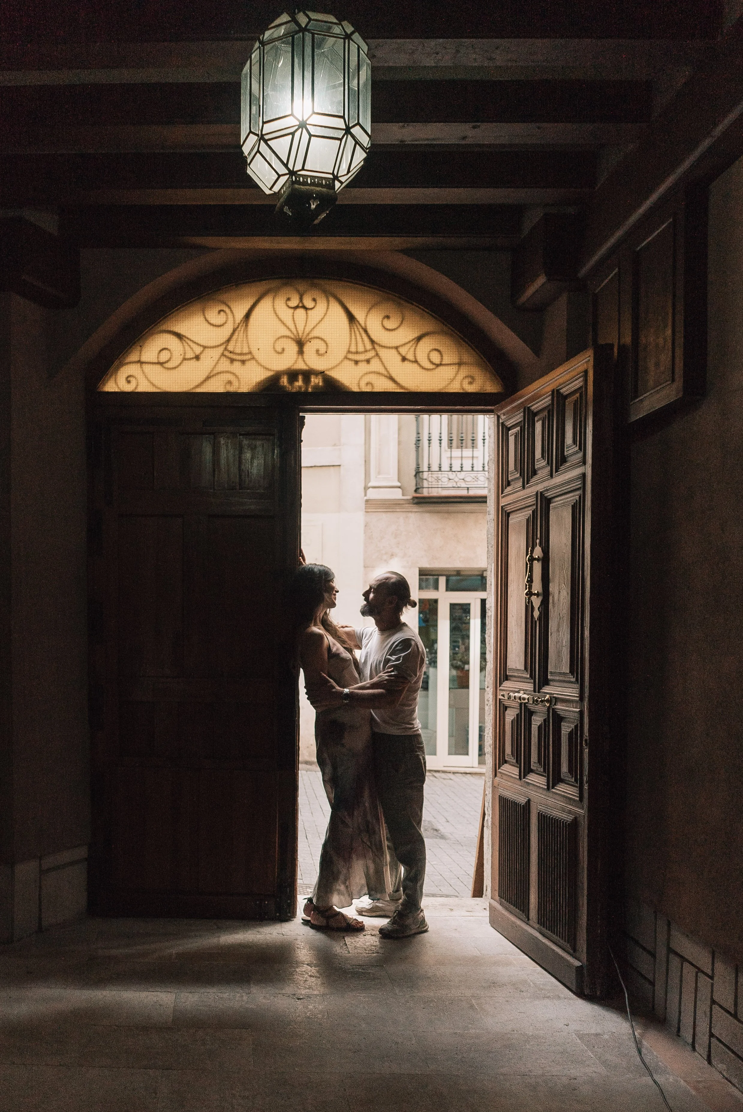 Casal sorridente de pé na entrada de uma residência, numa porta de madeira aberta, com luz natural vindo do exterior, formando um contraste com o interior escuro.