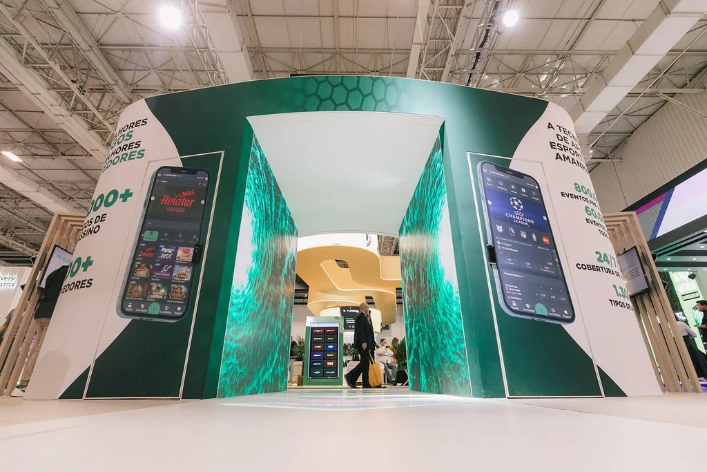 Estande de feira com paredes curvas decoradas com imagens de celulares que exibem aplicativos de apostas esportivas, com pessoas ao fundo.