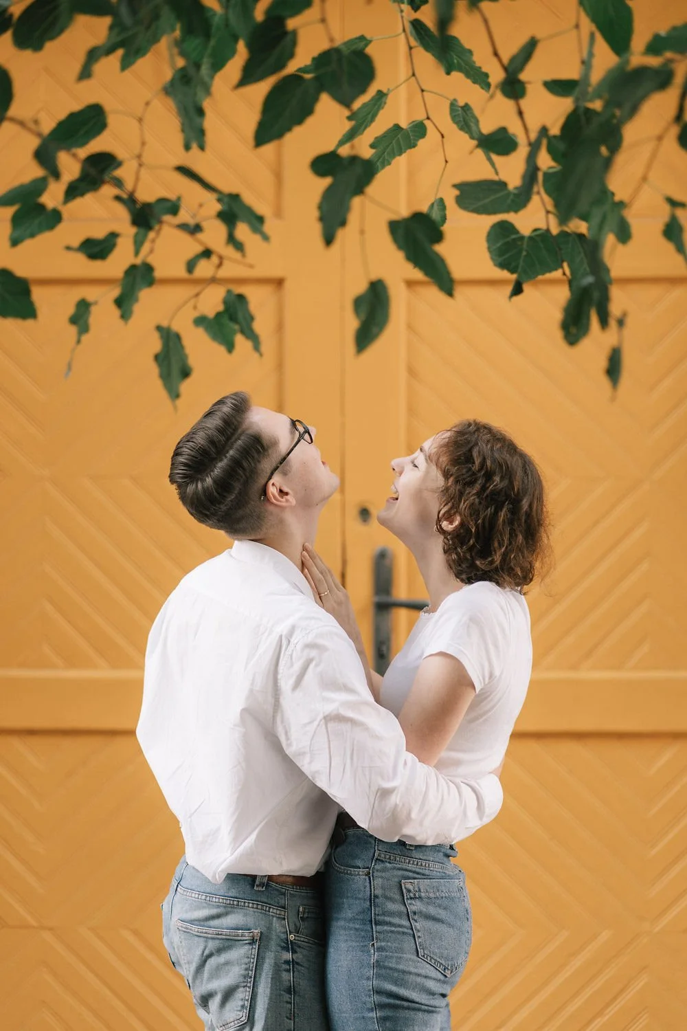 Um casal sorridente se abraçando de frente em um ambiente com fundo de madeira e vegetação.