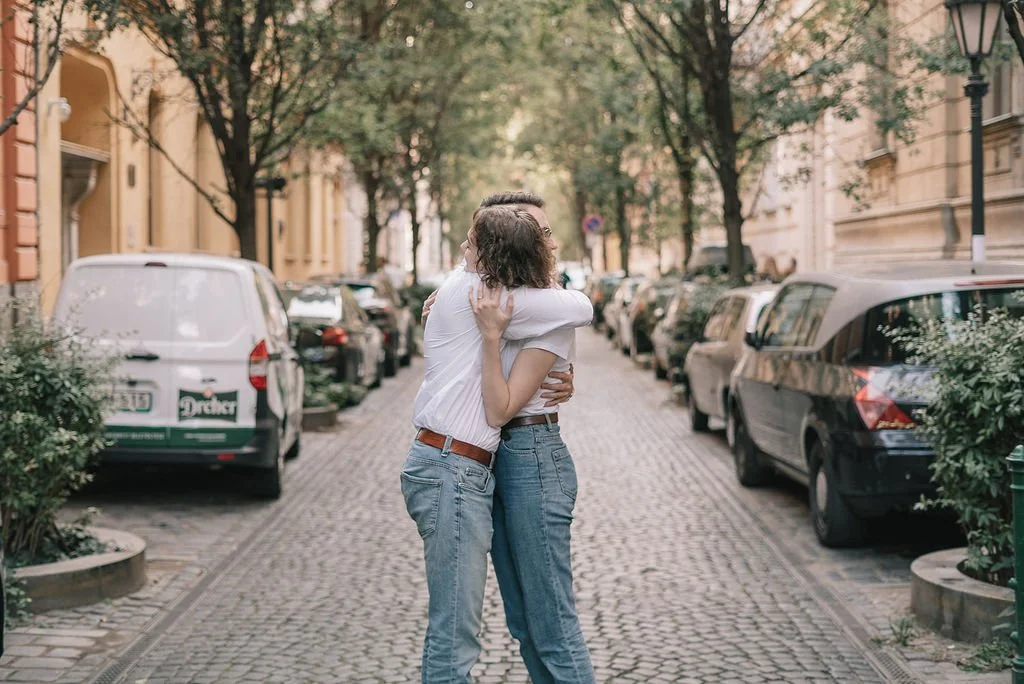 Dois jovens, um homem e uma mulher, se abraçando na rua, com carros estacionados ao lado e árvores ao fundo.