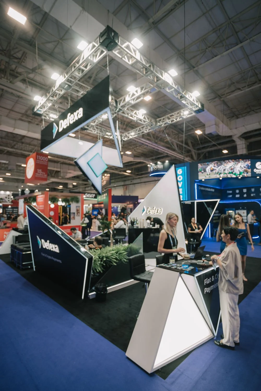 Feira ou evento com estandes tecnológicos da empresa Defexa, decoração moderna com painéis iluminados e pessoas conversando no expositor.