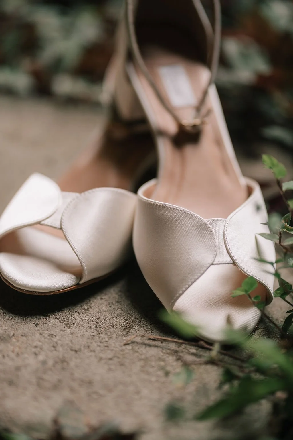 Sandálias de casamento brancas em monobloco com detalhes em fita, colocadas no chão ao lado de uma mochila de couro rosada.