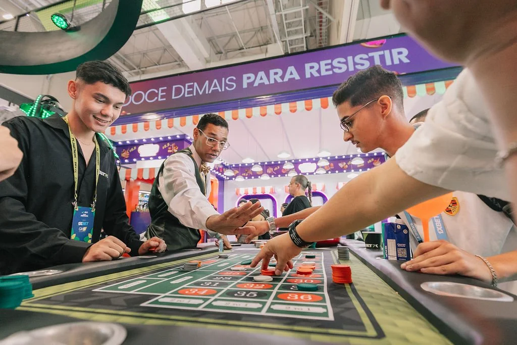 Pessoas jogando roleta em um cassino, com expositores ao redor e uma placa de fundo que diz 'DOCE DEMAIS PARA RESISTIR'.