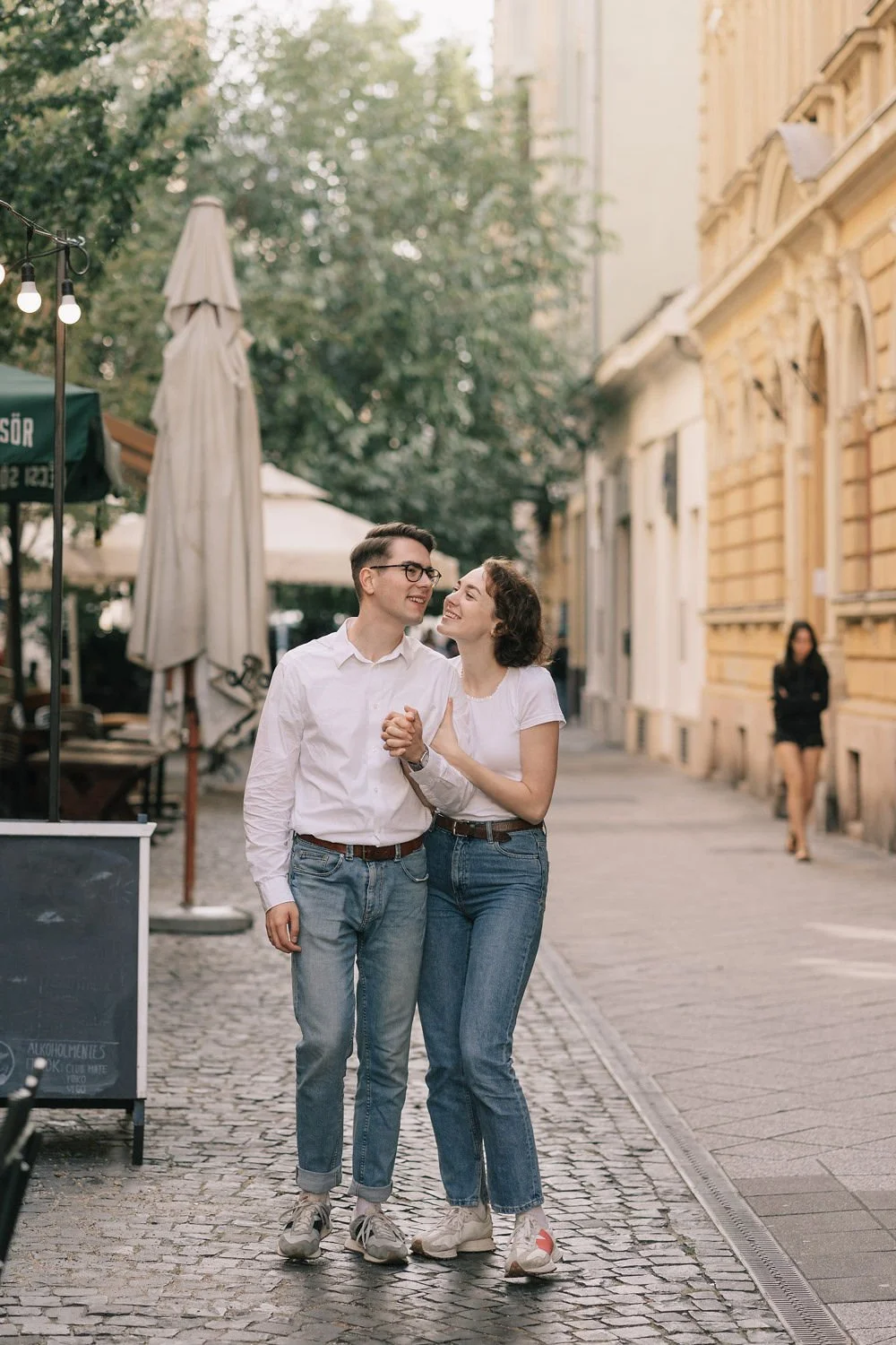 Casal sorridente caminhando de mãos dadas na rua de paralelepípedos, com construções antigas ao fundo e uma pessoa ao longe, em um dia ensolarado.