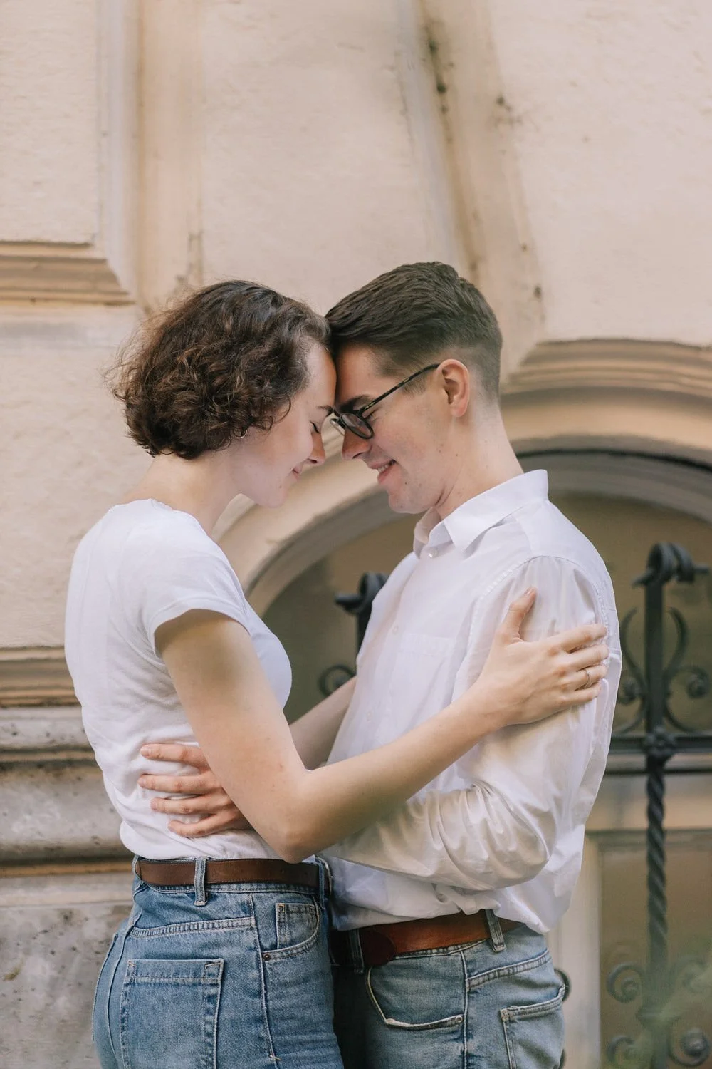 Casal jovem se abraçando com o nariz e testa tocando, sorrindo com os olhos fechados, diante de uma parede de pedra clássica.