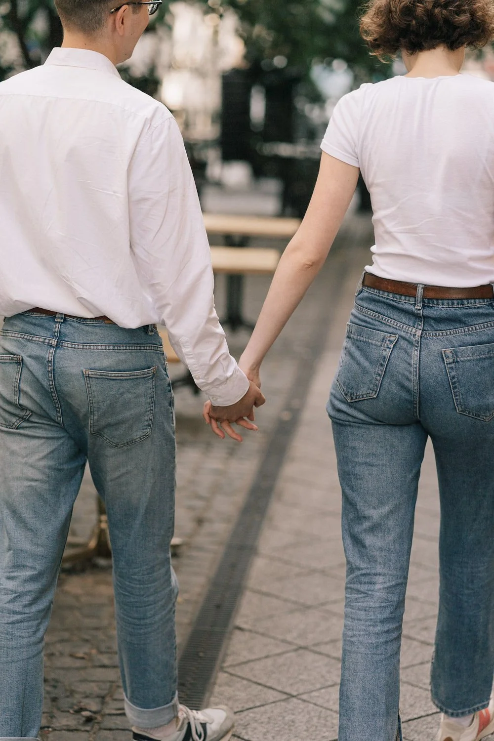 Casal de mãos dadas caminhando ao ar livre, vista de costas, ambos usando jeans e camisetas brancas.