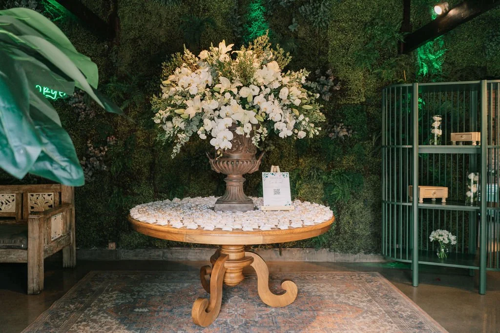 Arranjo de flores brancas em vaso decalcado sobre mesa de madeira com decoração de pétalas brancas ao redor, em ambiente com parede de plantas e iluminação suave.