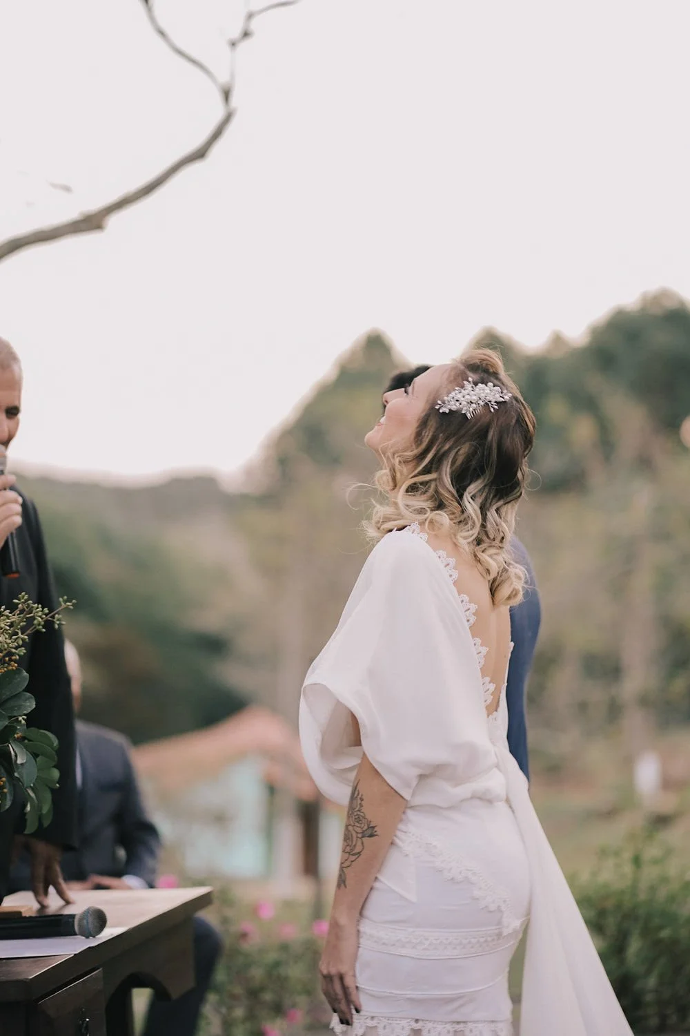 Noiva sorridente durante cerimônia ao ar livre, com cabelos cacheados e acessório de cabelo decorativo, usando vestido branco com detalhes em renda, ao lado de um homem vestido formalmente, em ambiente natural.