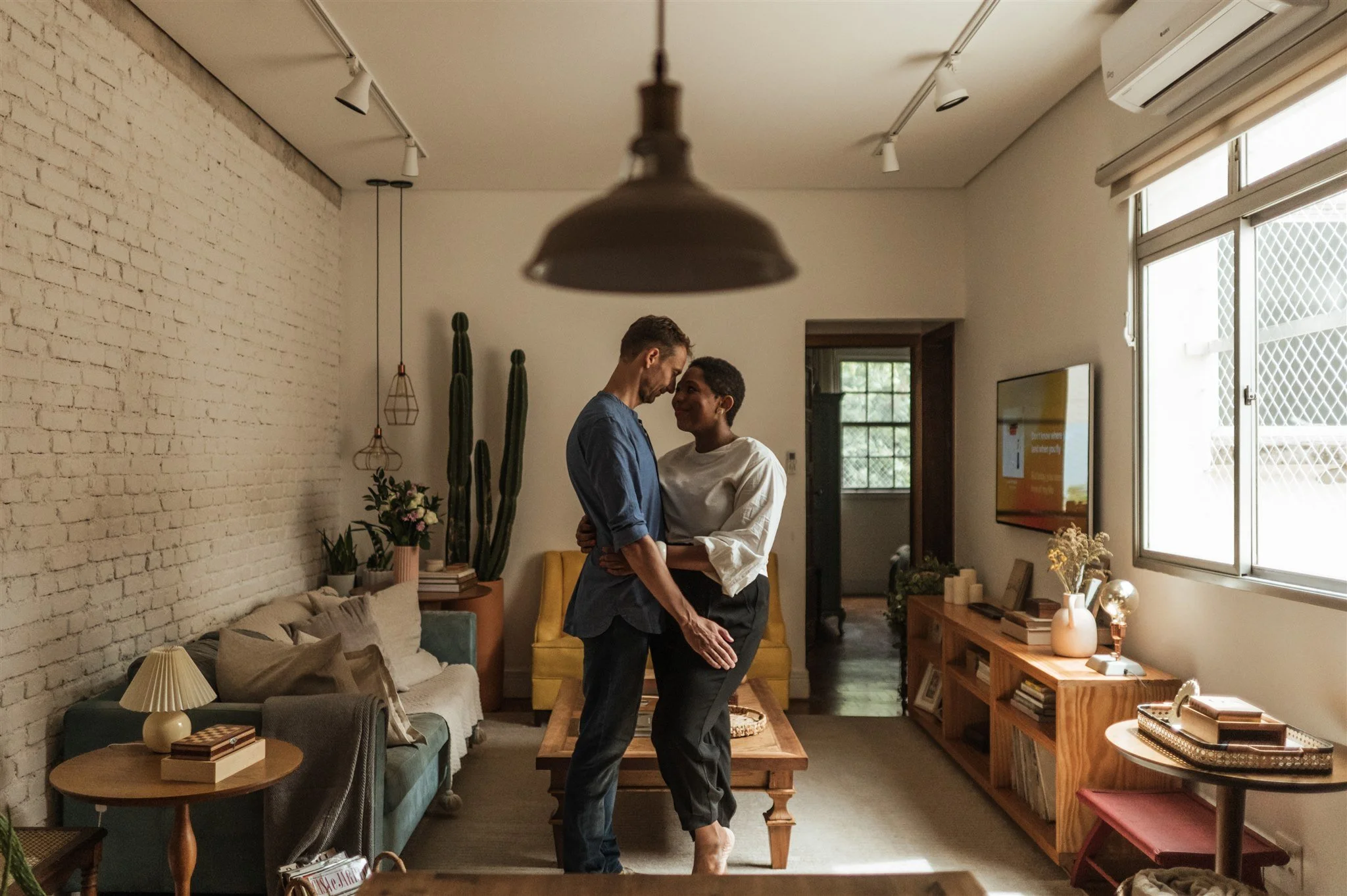 Couple embracing in cozy living room with brick walls, wooden furniture, and indoor plants.