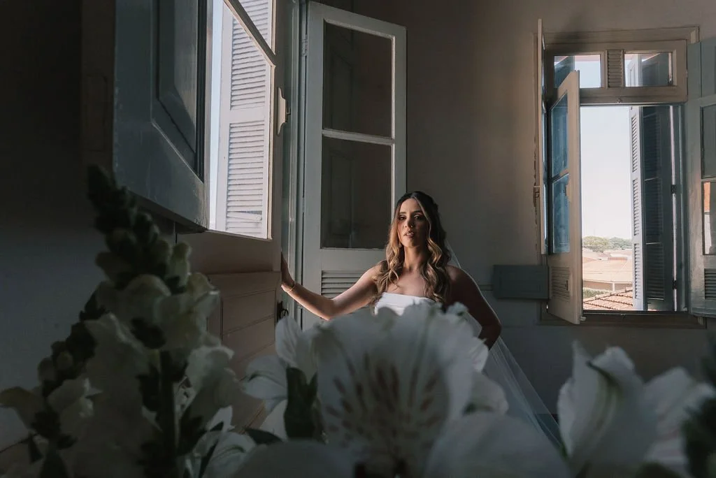 Mulher de vestido branco posando perto de janelas abertas em uma sala, com flores na frente.