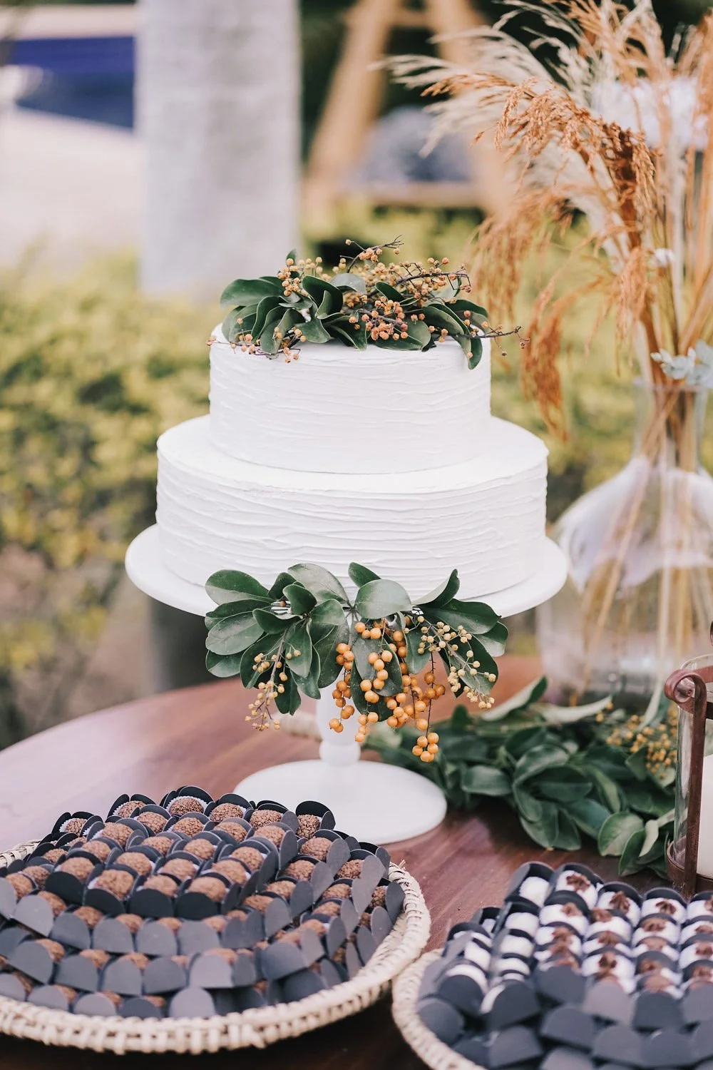 Bolo de casamento branco decorado com folhas verdes, amoras e sementes, colocado em suporte branco. Em volta, doces em forminhas pretas e brancas em cestas de palha. Fundo com vegetação e elementos decorativos rústicos.