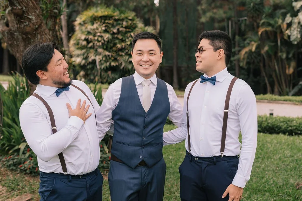 Três homens sorrindo e conversando ao ar livre, dois deles vestindo camisa branca, suspensórios e gravata borboleta, e um no centro usando terno azul com colete, em um jardim com plantas e árvores ao fundo.