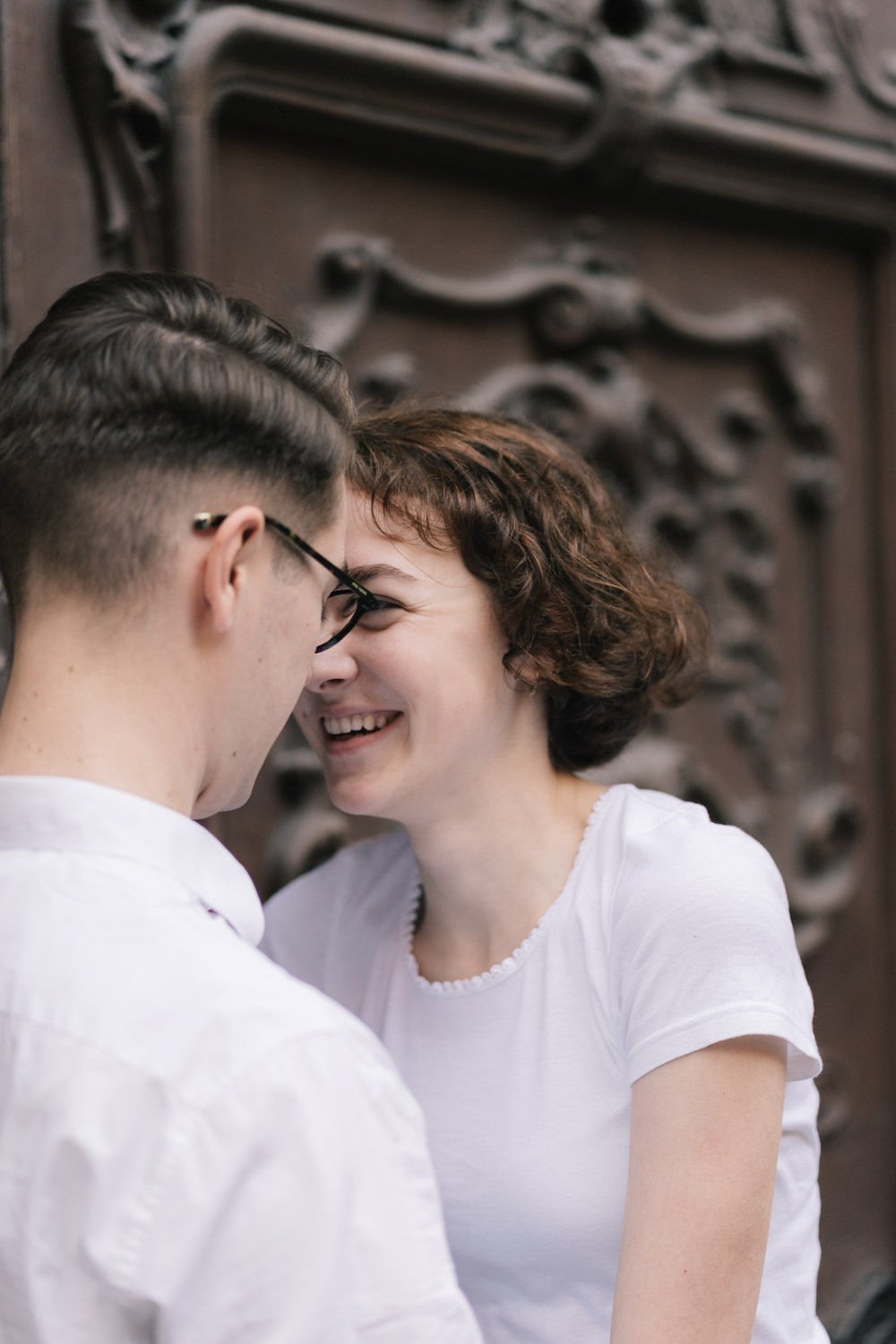 Jovem casal brincando e sorrindo com a testa e os rostos quase tocando, em um ambiente decorado com porta de madeira entalhada.