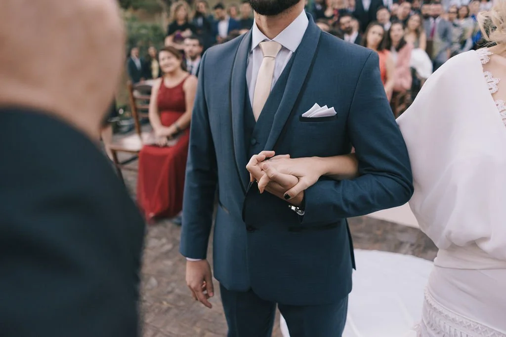 Casal de casamento trocando alianças ao ar livre, com convidados ao fundo.