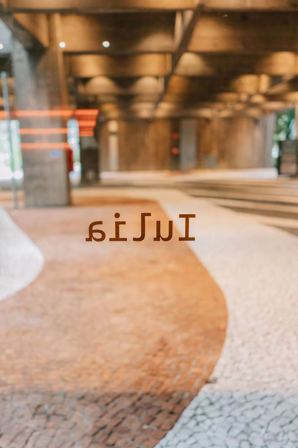 Letra espelhada na porta de vidro que diz 'Itaú', com interior do banco visível ao fundo, incluindo piso de pedras e elementos de madeira.