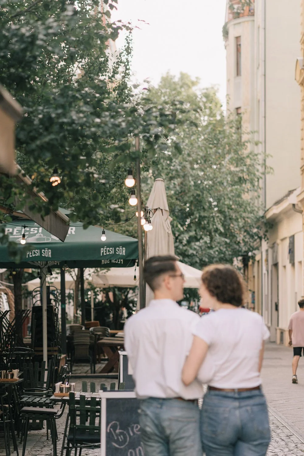 Casal de jovens andando de mãos dadas na rua de uma cidade, ao entardecer, com árvores, mesas de um restaurante ao ar livre, e edifícios altos ao fundo.