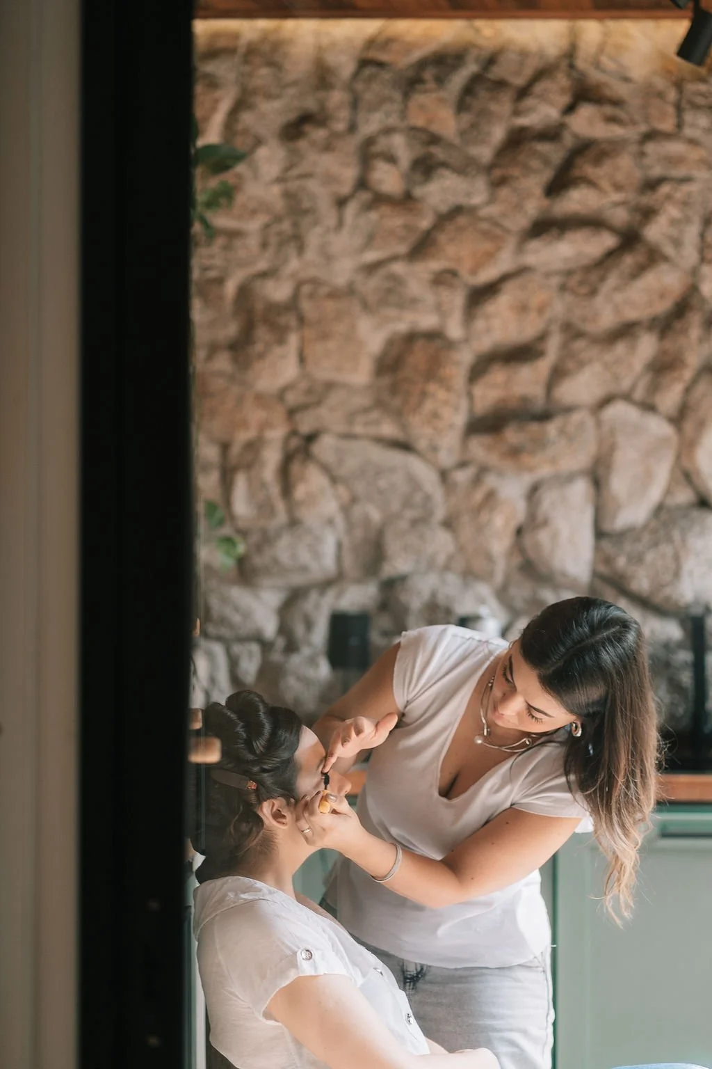 Uma mulher fazendo maquiagem em uma menina, com uma parede de pedras ao fundo e uma iluminação suave.