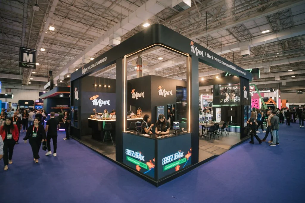 Estande de feira com nome 'Moen', decorado com iluminação moderna e várias pessoas interagindo, em um evento de grande porte.