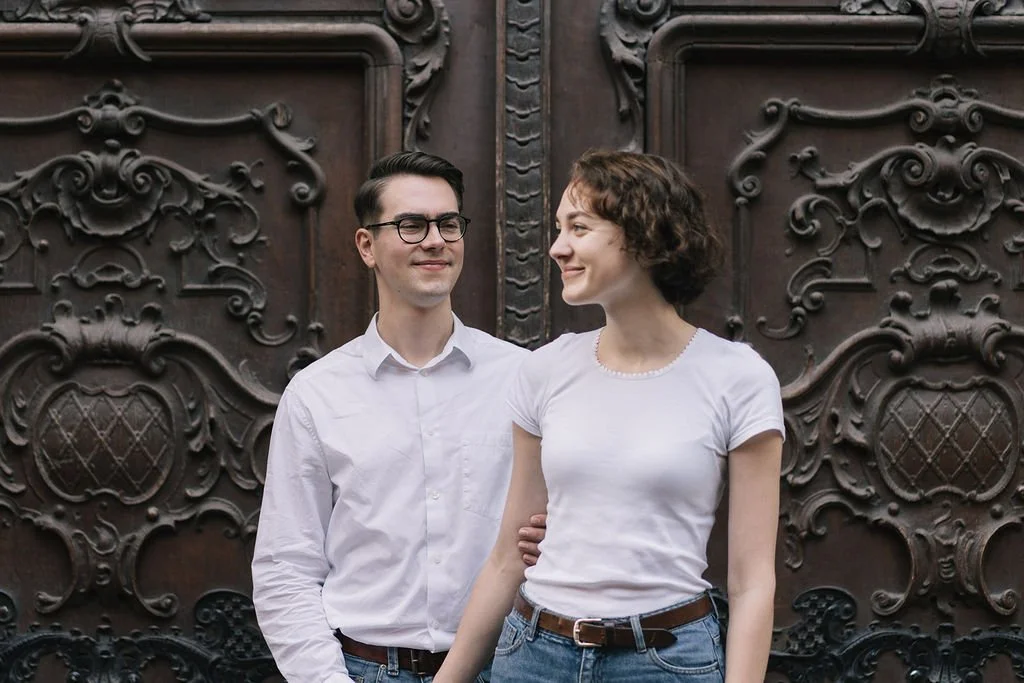 Homem e mulher sorrindo, de pé juntos na frente de uma porta de ferro ornamentada.