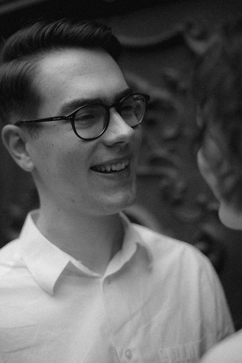 Homem sorrindo usando óculos de grau, em preto e branco, aparência elegante, fundo desfocado.