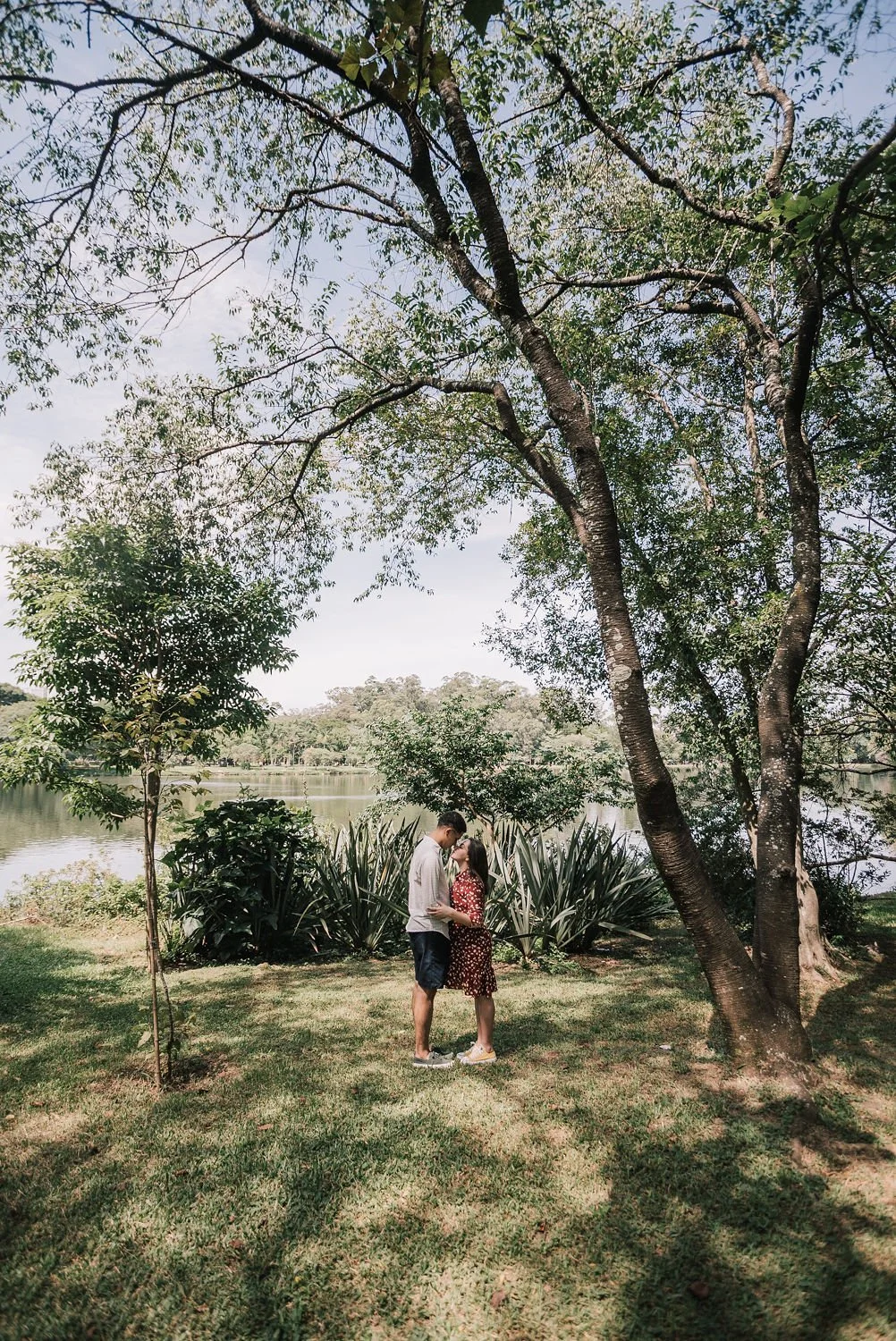 Um casal se beijando ao lado de um rio, cercado por árvores e vegetação, sob um céu claro.