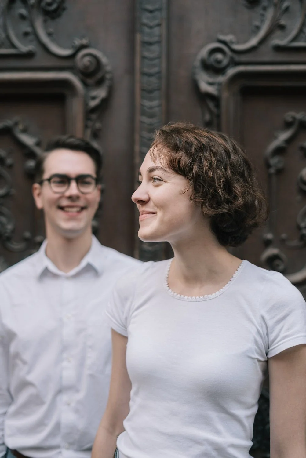 Duas pessoas sorrindo, uma mulher ao centro e um homem ao fundo, ao lado de uma porta de madeira com detalhes esculpidos.