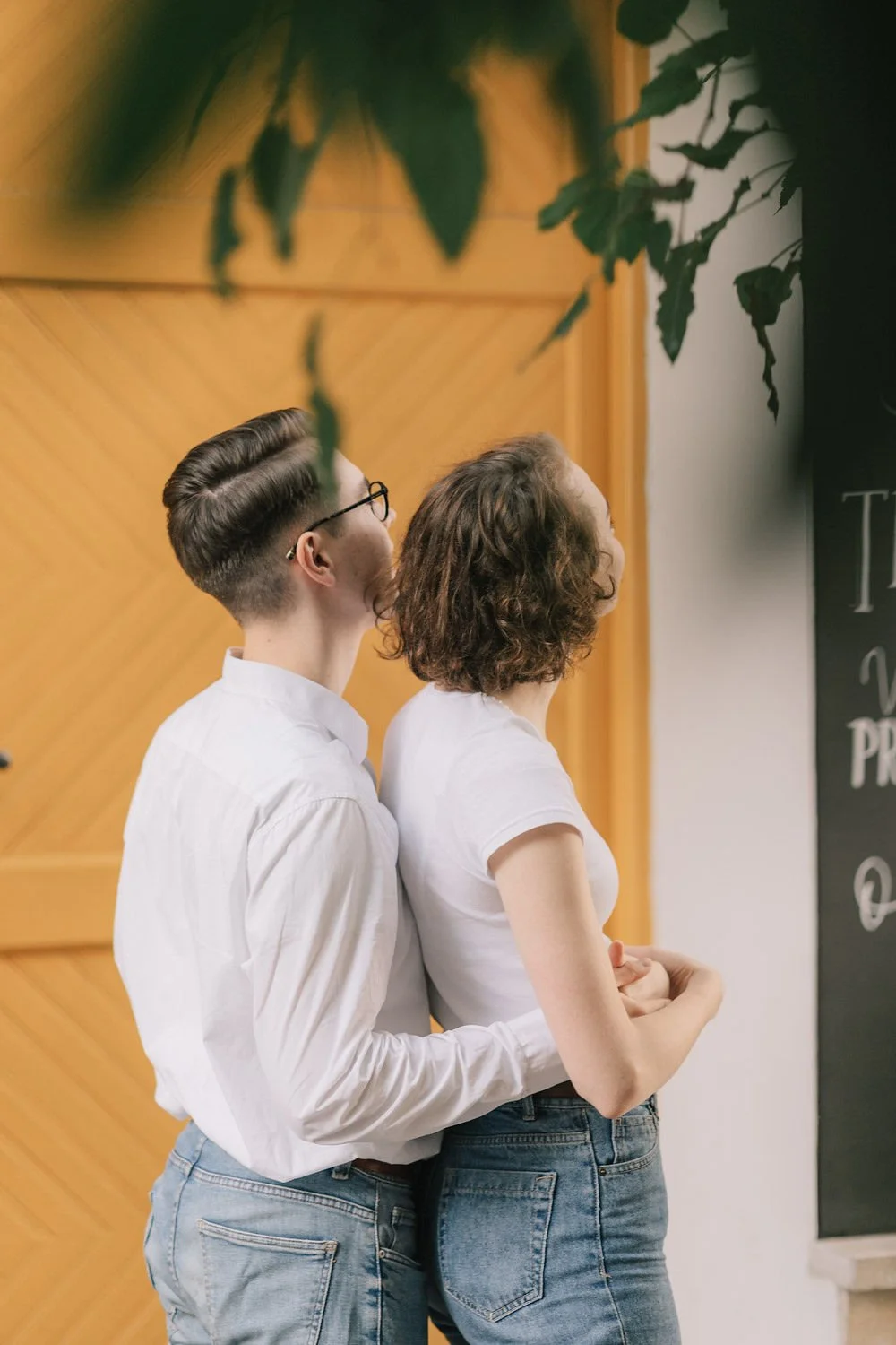 Homem e mulher de costas, juntos, em uma fila ou sala, com fundo de parede de madeira, vestindo camisetas brancas e jeans, com folhas de árvores visíveis ao fundo.